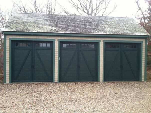 A three-bay detached garage with dark green carriage-style doors and tan trim, set against a backdrop of bare trees.
