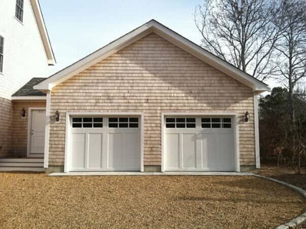 A two-car garage with white doors and wood shingle siding attached to a white house, seen from a gravel driveway.