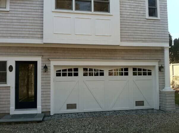 A white garage with windows along the top and a black-framed side door, set against a light grey shingled house exterior.