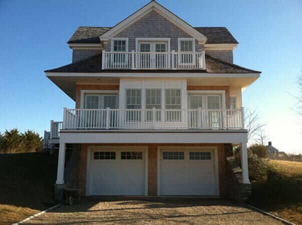 A three-story, shingled coastal house with a double garage, white railings, and balconies under a clear blue sky.