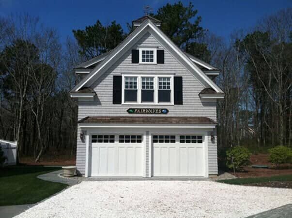 A two-story, gray shingled carriage house with a white double garage, black shutters, and a sign reading 