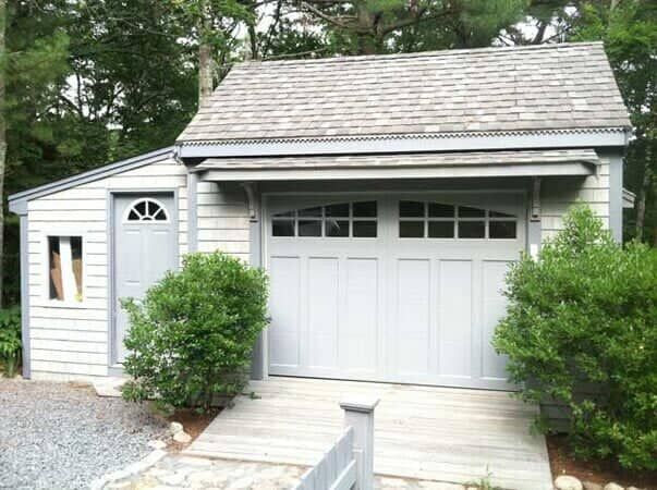 A light-gray detached garage with a wooden deck entrance, a side door, and a single-car overhead door in a wooded area.