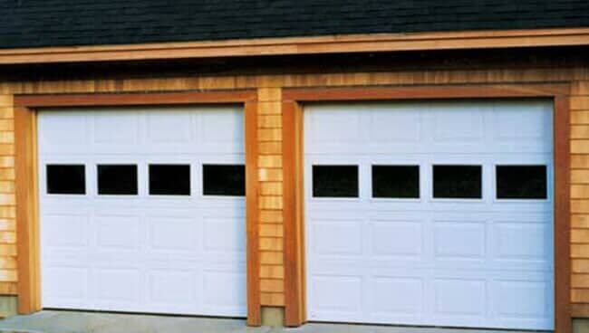 Two white garage doors with four rectangular window panels each, set into a building with light brown cedar shake siding.