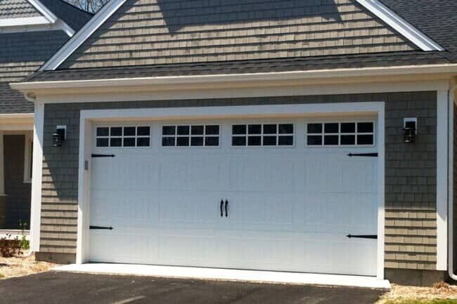 White single-car garage door with decorative windows and black hardware on a gray shingle-sided house.