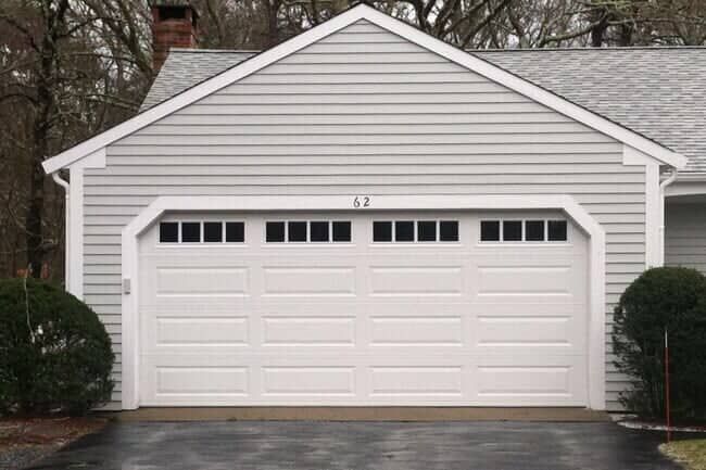 A single-story residential garage with grey horizontal siding, a white door featuring window panels, and a paved driveway.