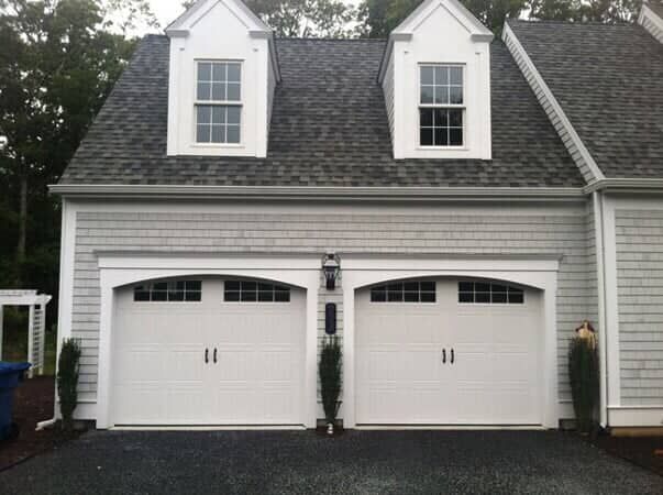 Exterior of a two-car garage with arched white doors, gray shingle siding, and two dormer windows on the dark roof.