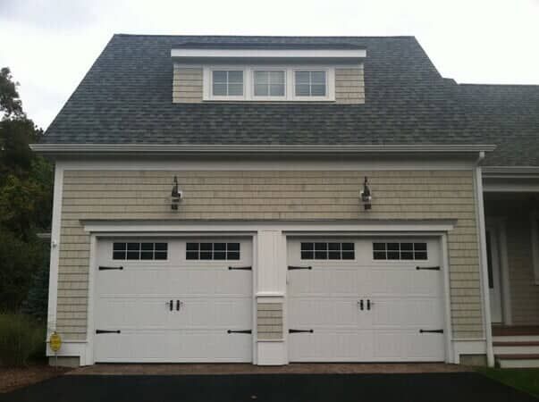 Two white carriage-style garage doors with windows, set in a light beige shingled building with a small roof dormer.