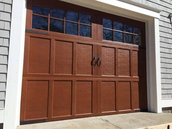 A brown, carriage-style garage door with glass windows, installed on a house with gray shingles.