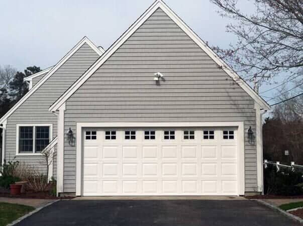A gray-sided house with a white two-car garage door and a window, viewed from a paved driveway.