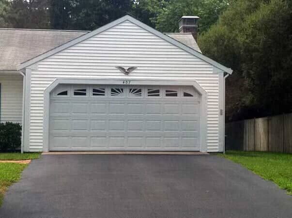 White garage with a sunburst window top and bird decoration, featuring a gray asphalt driveway and lush green surroundings.