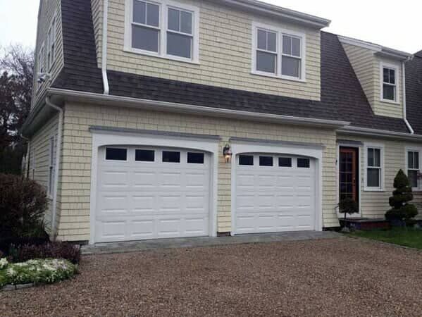 A two-story house with beige cedar shake siding, dark roof shingles, and two white garage doors with arched windows.