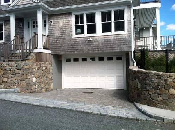 A shingled house with a white garage door, surrounded by stone retaining walls and a paved driveway.