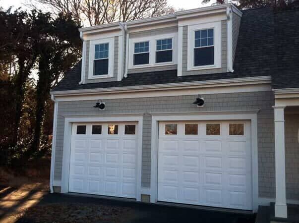A two-car garage with white doors and a dormer featuring three windows set into gray shingled siding.