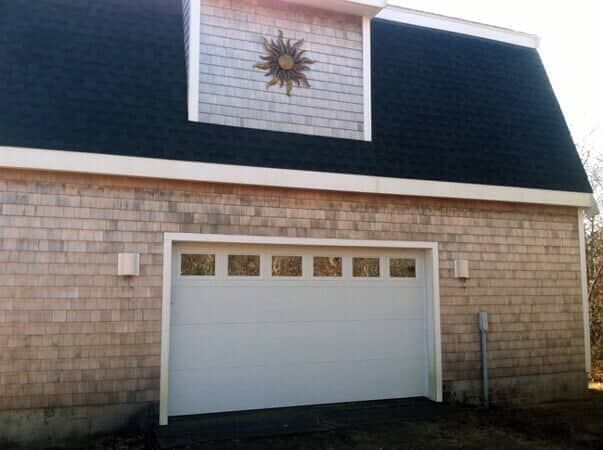 A gray shingled garage with a black gambrel roof, a white garage door, and a metal sun decoration above the door.