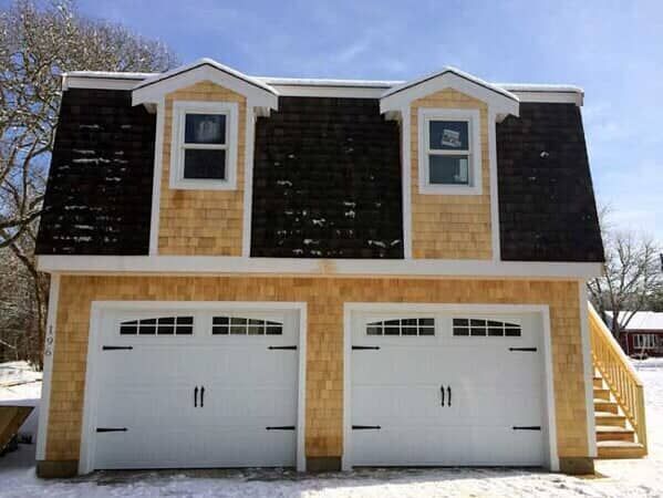 A two-car garage with light wood shingles, two white carriage-style doors, a gambrel roof, two dormer windows, and stairs.