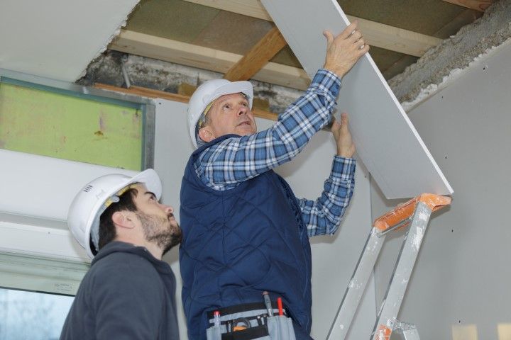 Two workers installing a gray ceiling panel on a ladder indoors