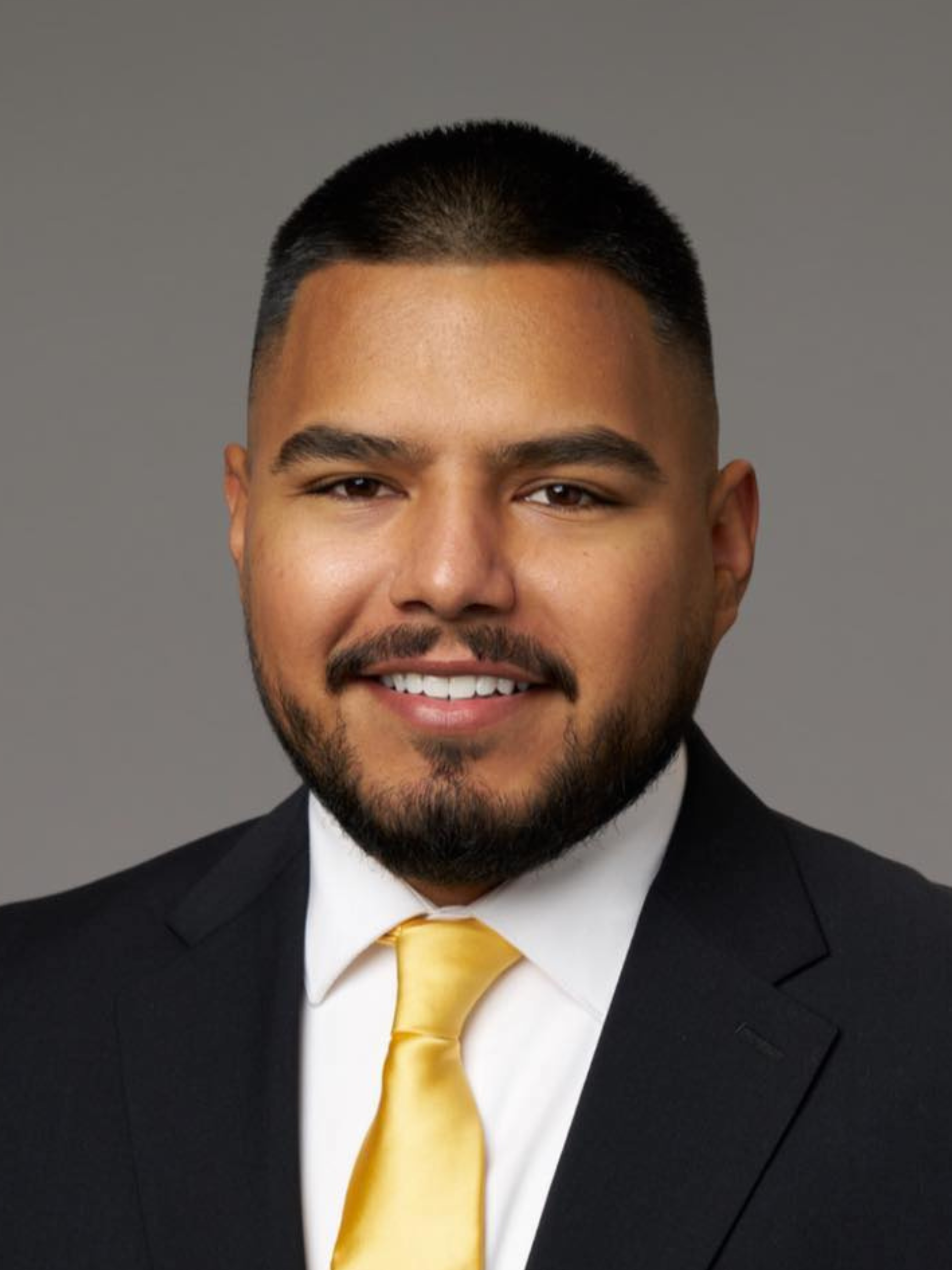 Headshot of a smiling person with a beard, wearing a dark suit jacket, white shirt, and yellow tie against a grey backdrop.