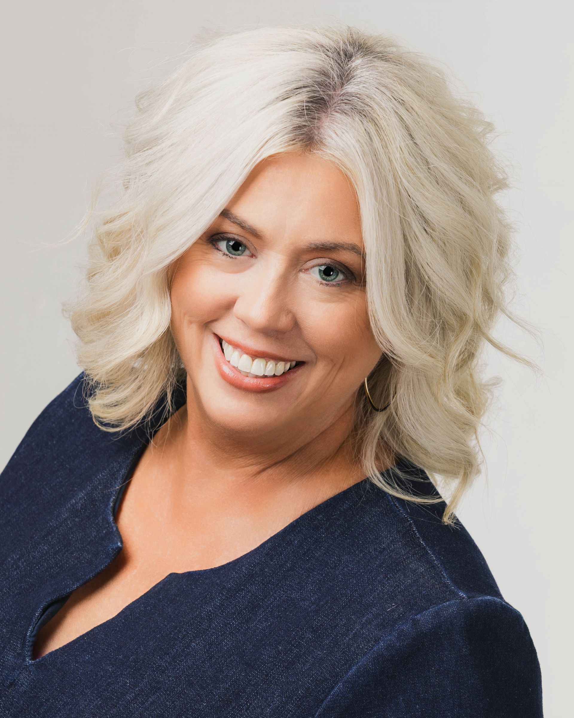 Headshot of a smiling person with shoulder-length, wavy, light-colored hair, wearing a textured dark blue blazer.