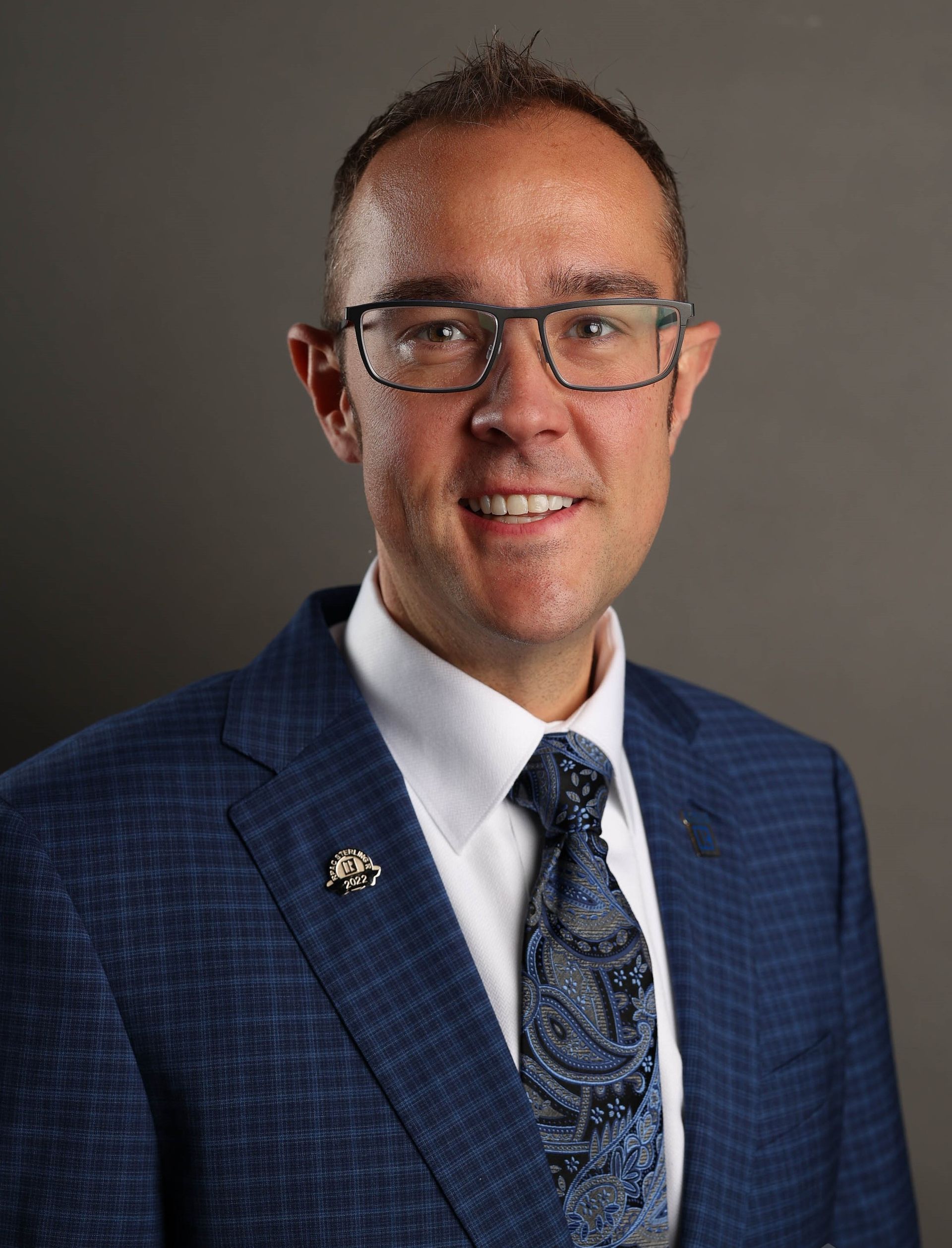 A person wearing a patterned blue suit, white shirt, patterned tie, and glasses, smiling against a dark gray background.