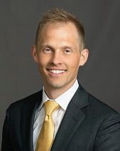 A professional headshot of a person wearing a dark suit, white shirt, and yellow tie, smiling against a gray background.