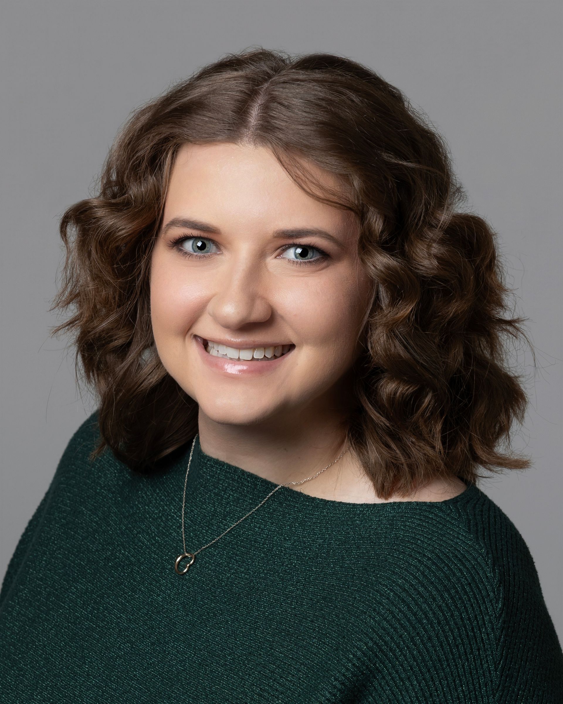 A smiling person with wavy brown hair wearing a black top and a small heart necklace against a gray background.