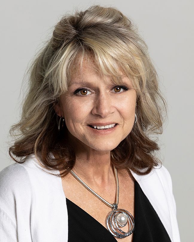 A smiling woman with blonde hair, wearing a white cardigan, a black top, and a circular pendant necklace.
