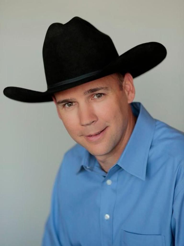 A man in a blue button-down shirt and a black cowboy hat smiling against a plain, light gray background.