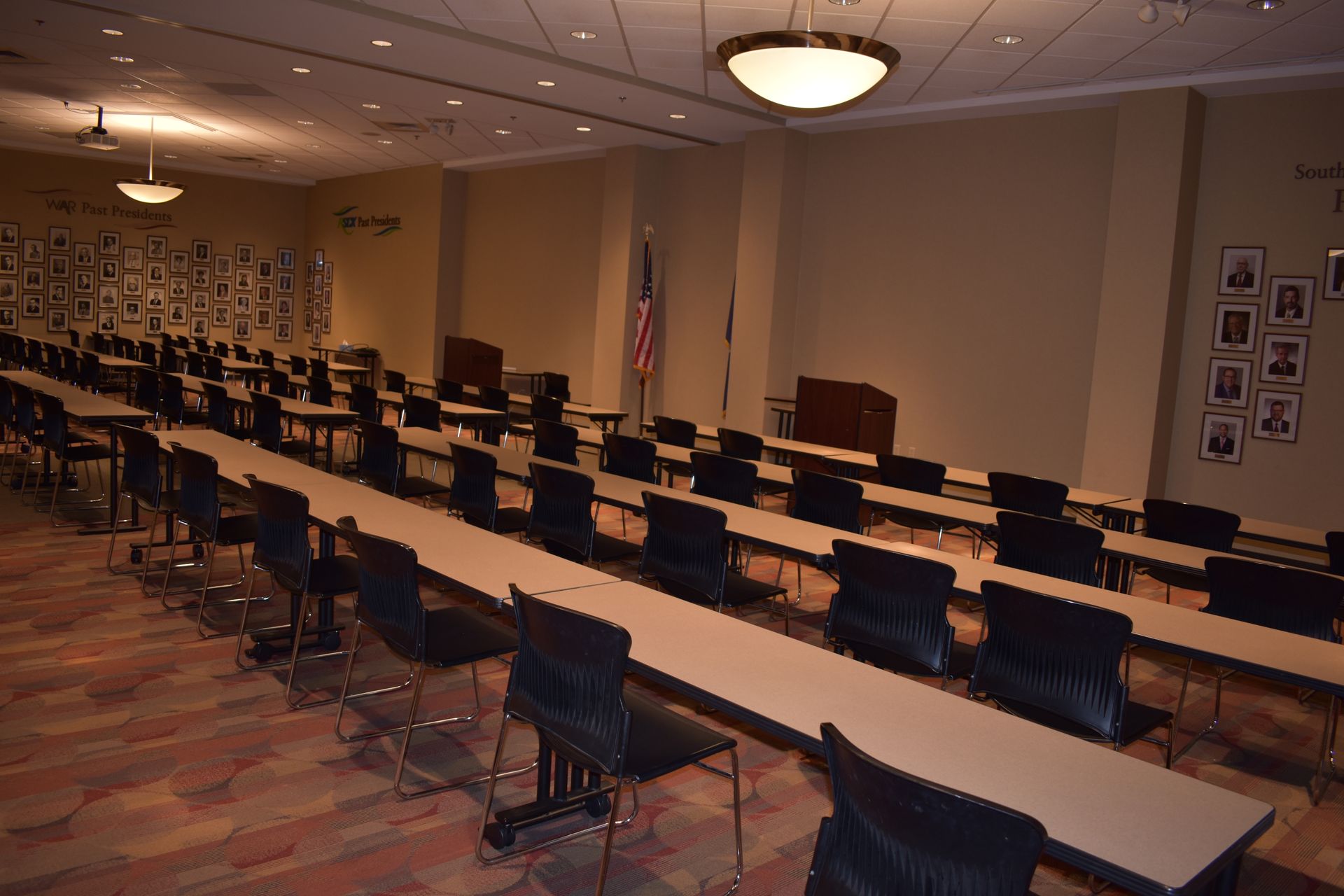 An empty classroom or meeting hall with rows of tables and chairs, wood-paneled walls, and displays of framed photos.