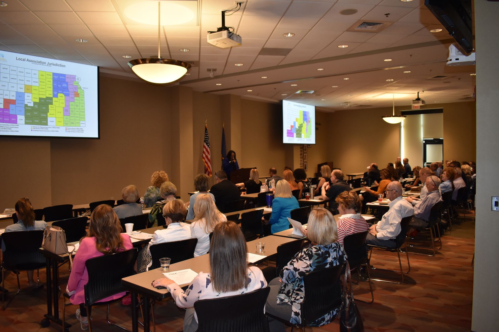 A speaker addresses an audience seated at tables in a conference room with maps projected on screens at the front.
