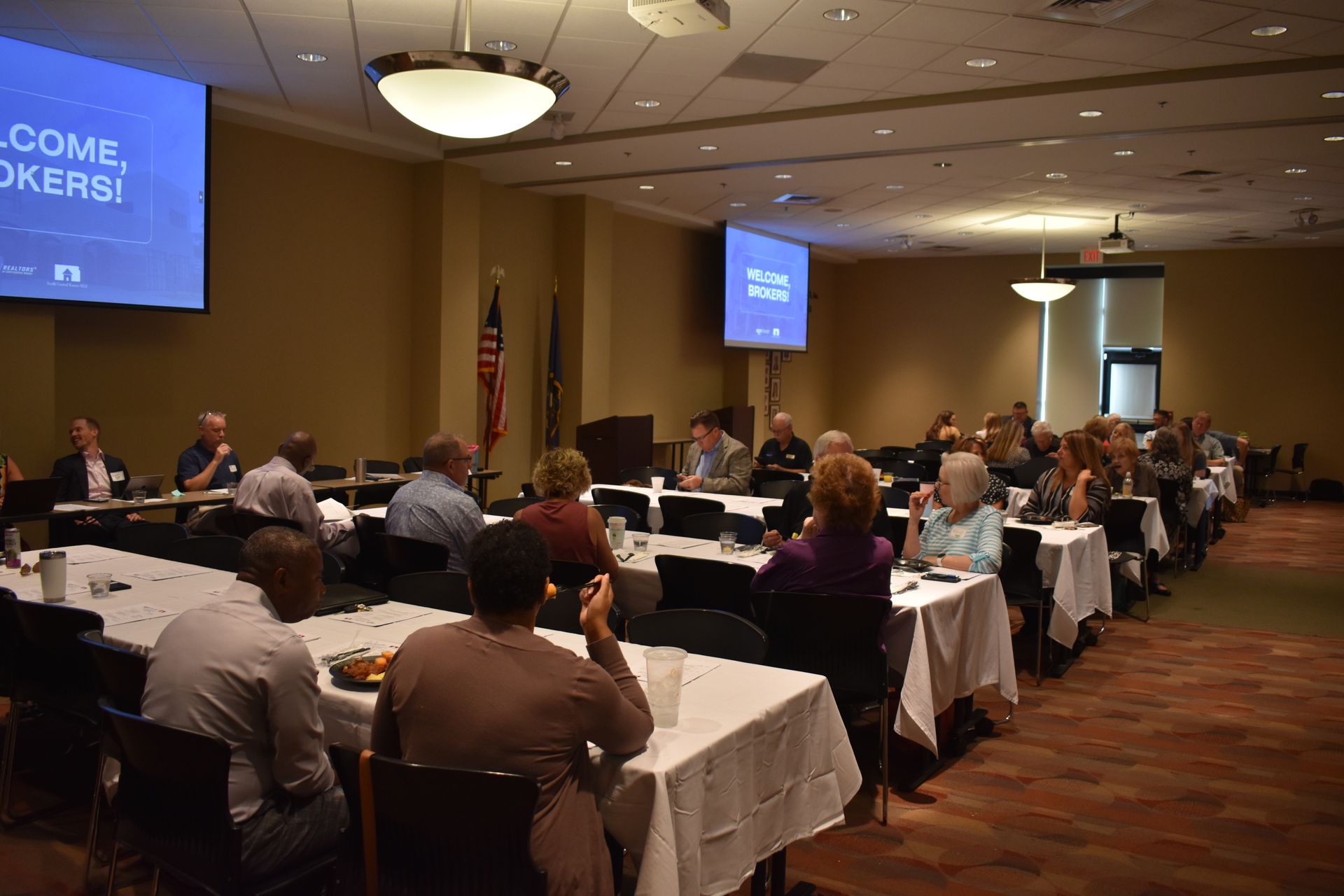 A group of people seated at tables in a conference room attending a presentation with slides projected on screens.