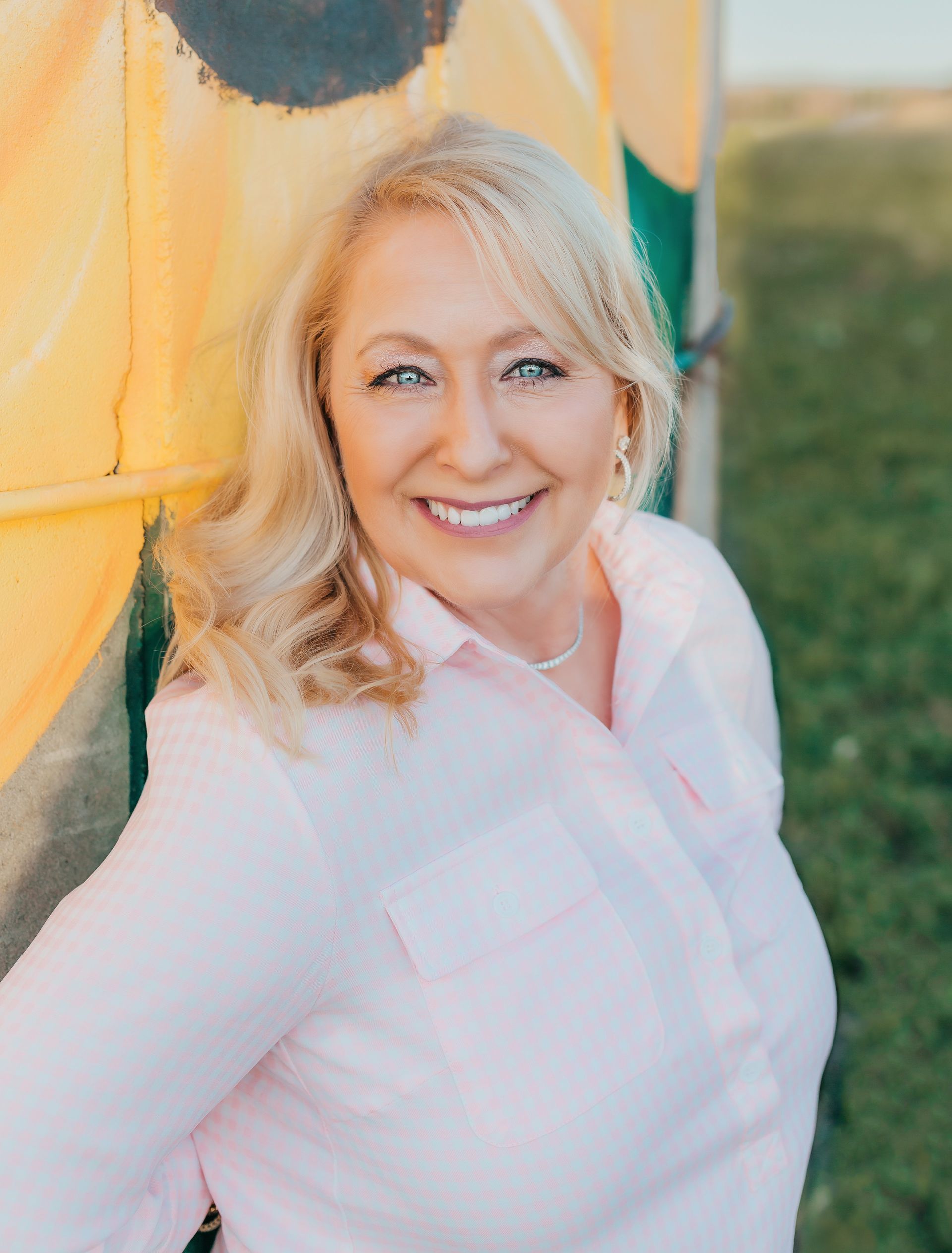A smiling person with blonde hair wearing a light pink button-down shirt, standing outdoors against a yellow background.
