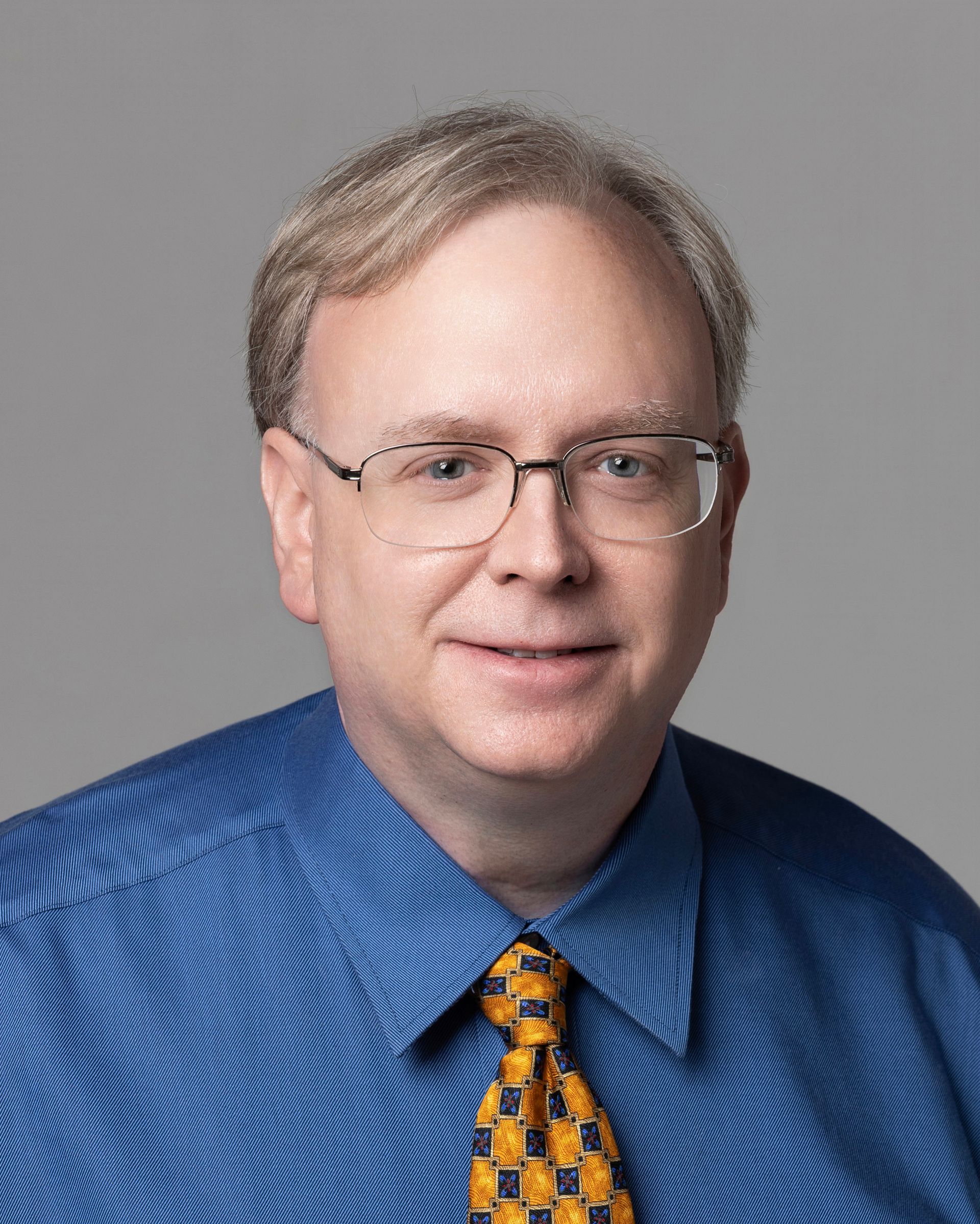 A professional headshot of a person wearing glasses, a white dress shirt, and a patterned tie against a gray background.