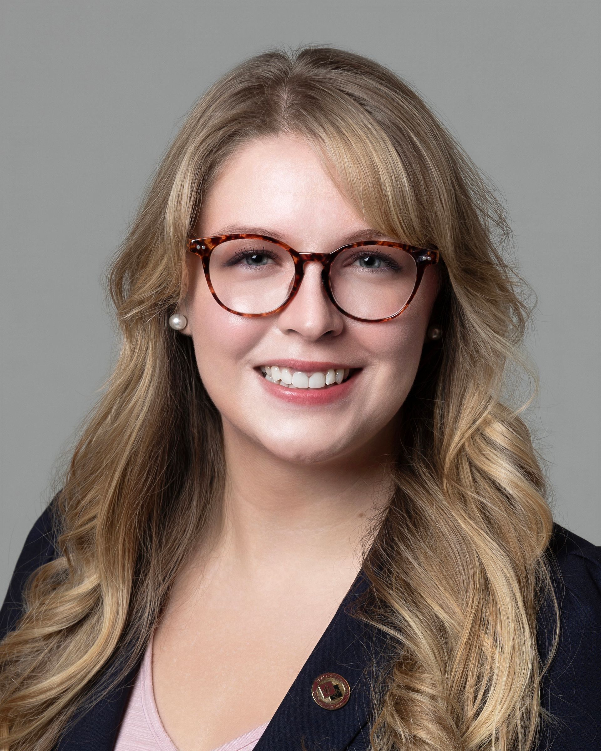 A headshot of a person wearing glasses and a gold chain necklace, smiling against a plain gray background.