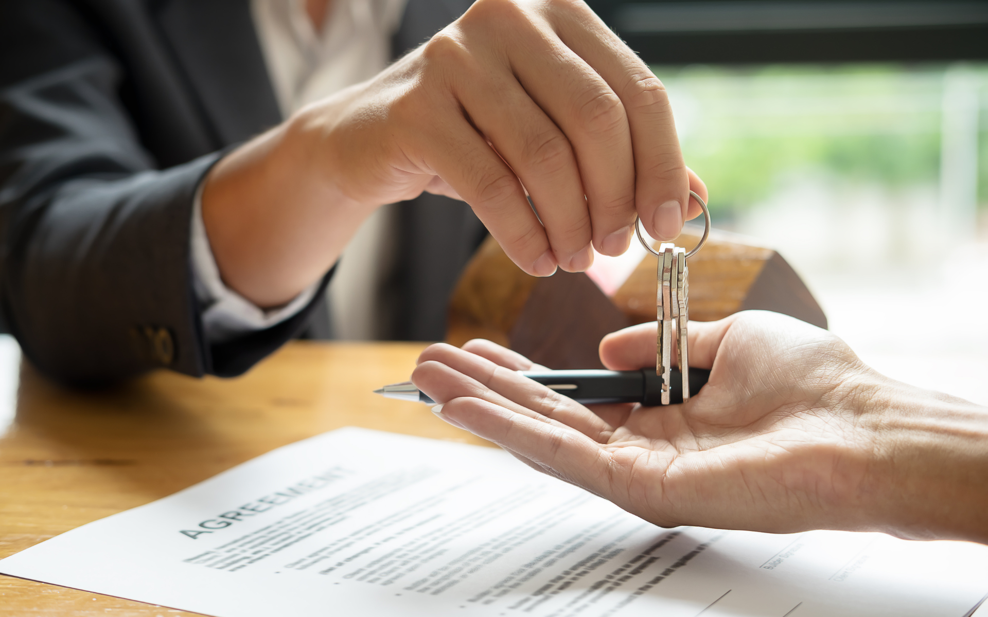 A person in a suit hands house keys and a pen to another person over a real estate contract on a wooden desk.