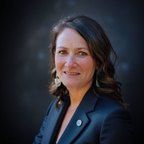 Headshot of a smiling person wearing a dark blazer with a small lapel pin against a dark, mottled background.