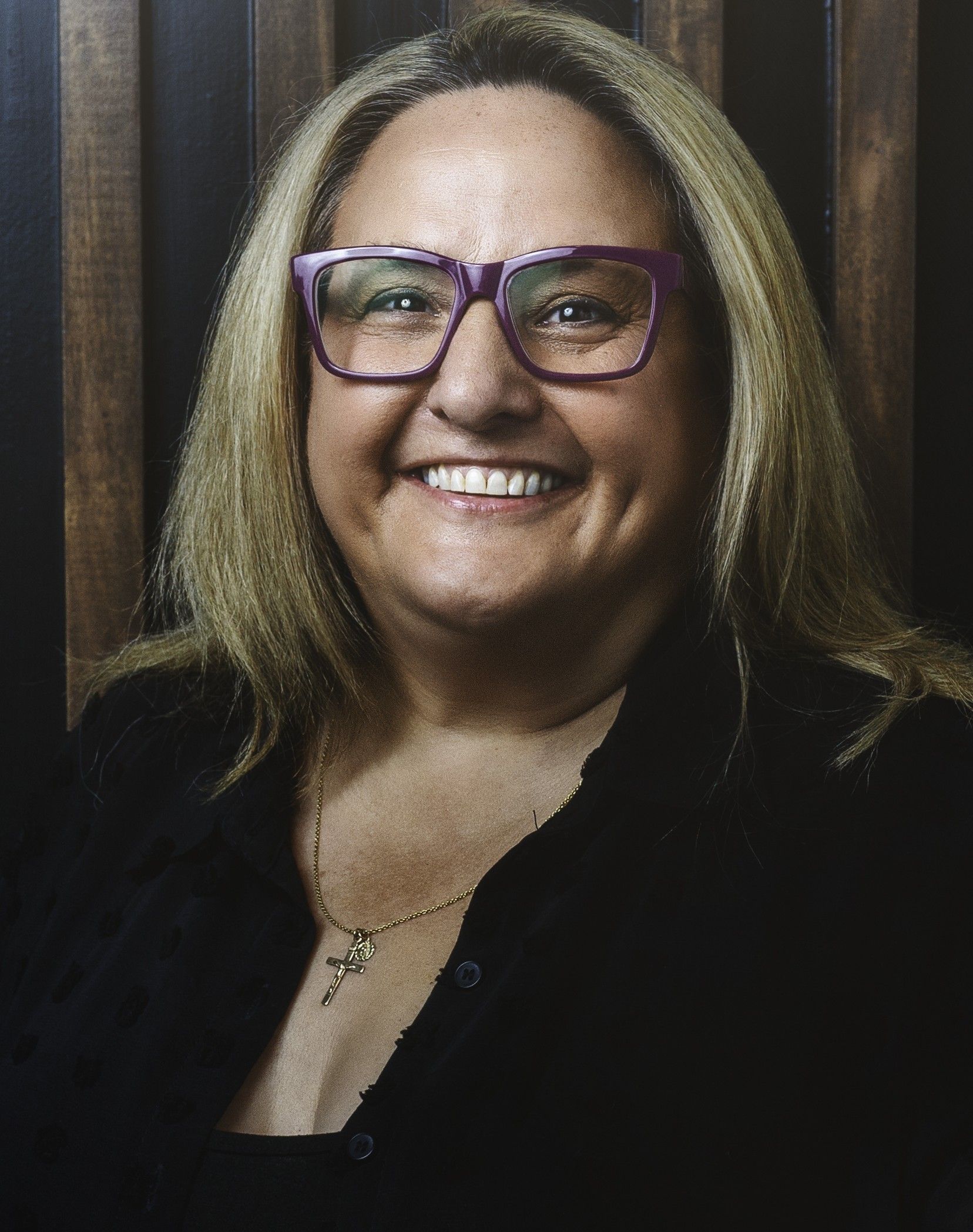 A smiling person with shoulder-length blonde hair, wearing a black turtleneck, posed against a solid grey background.