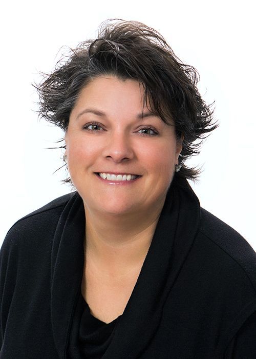 A professional headshot of a smiling person with dark, layered hair wearing a black top against a white background.