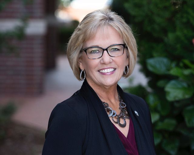 Headshot of a smiling person with blonde hair and glasses, wearing a black blazer and a necklace, posed outdoors.