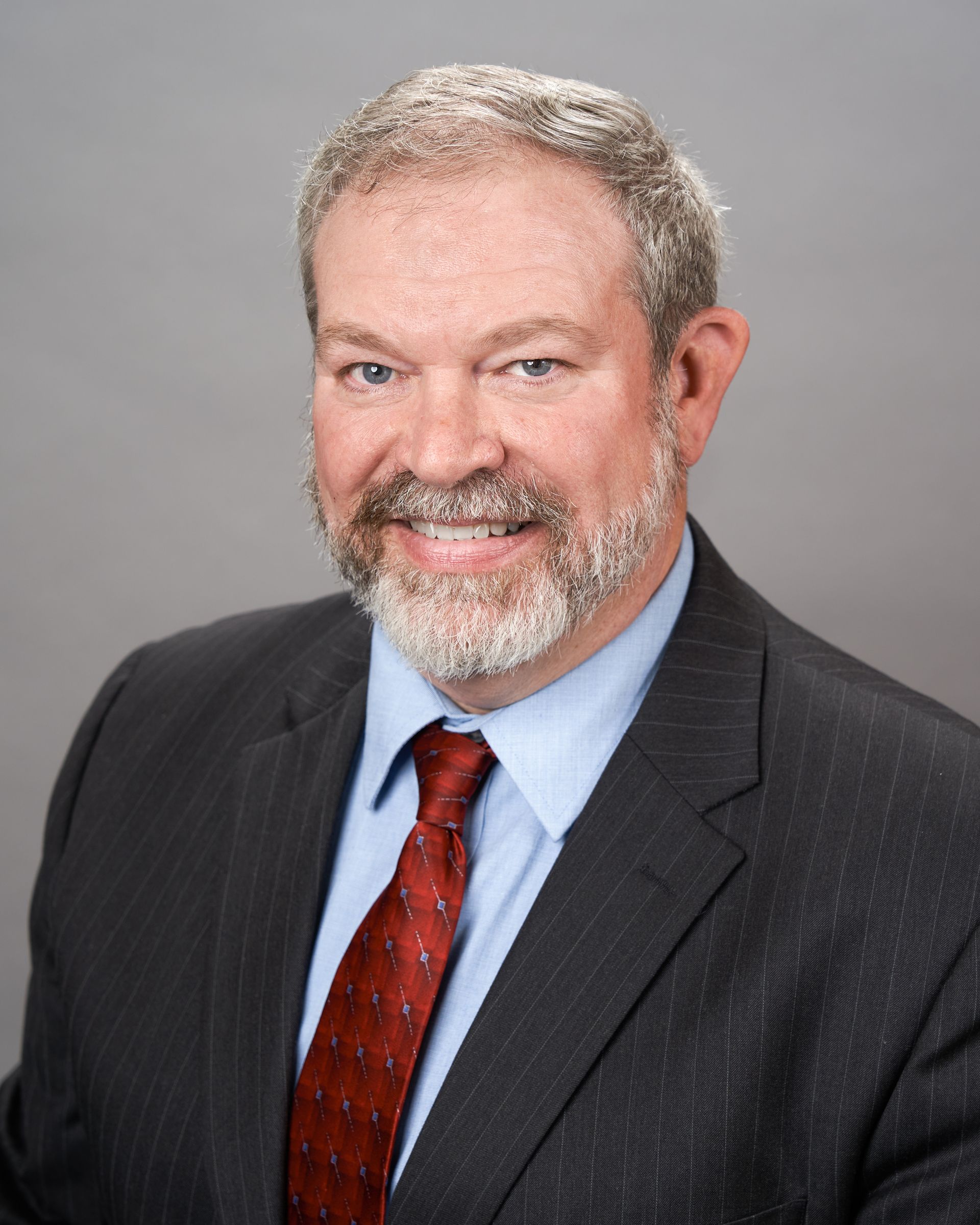 A professional headshot of a smiling man with a beard, wearing a pinstripe suit, light blue shirt, and red patterned tie.