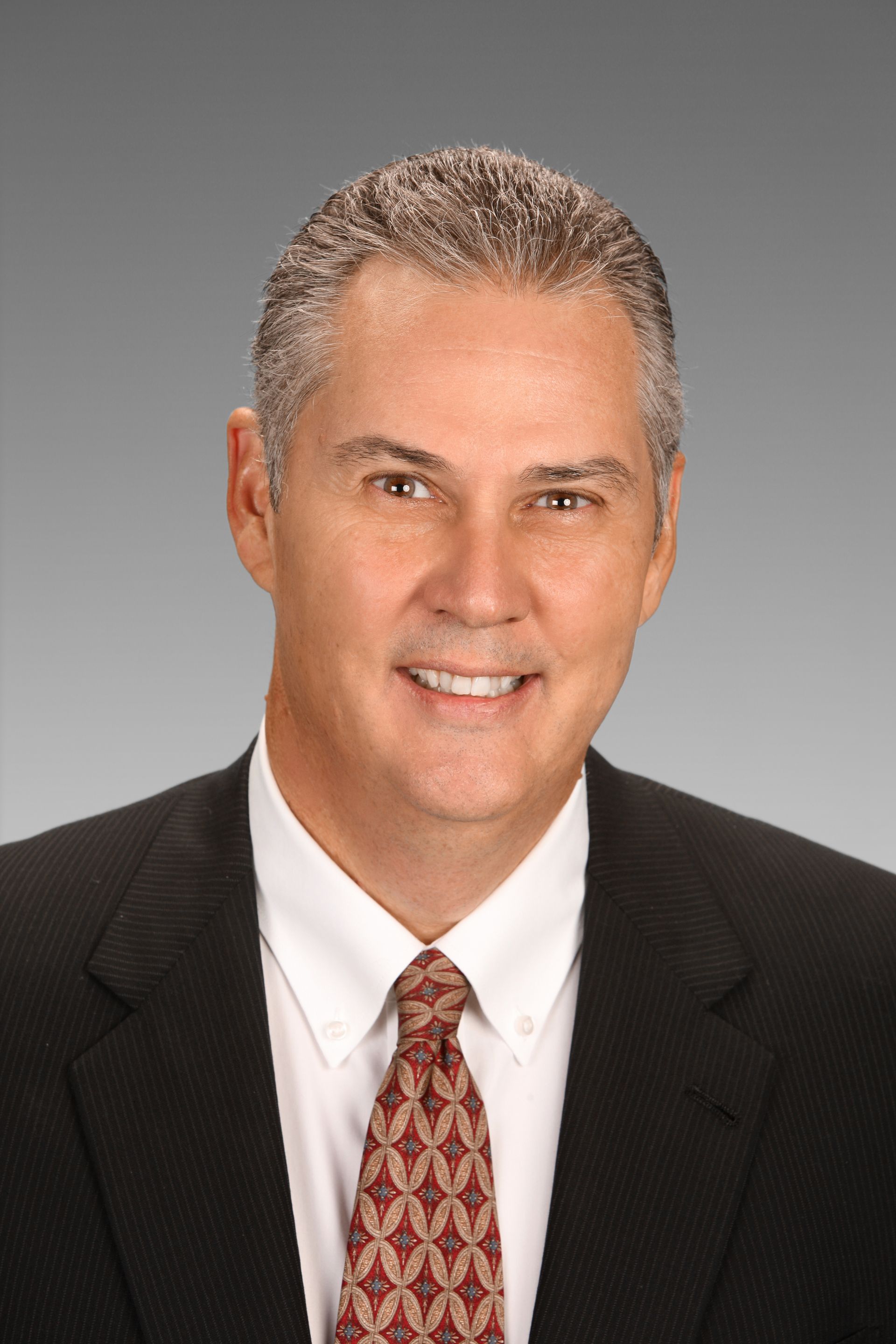 A professional headshot of a smiling man wearing a black suit, white shirt, and patterned red tie against a gray background.
