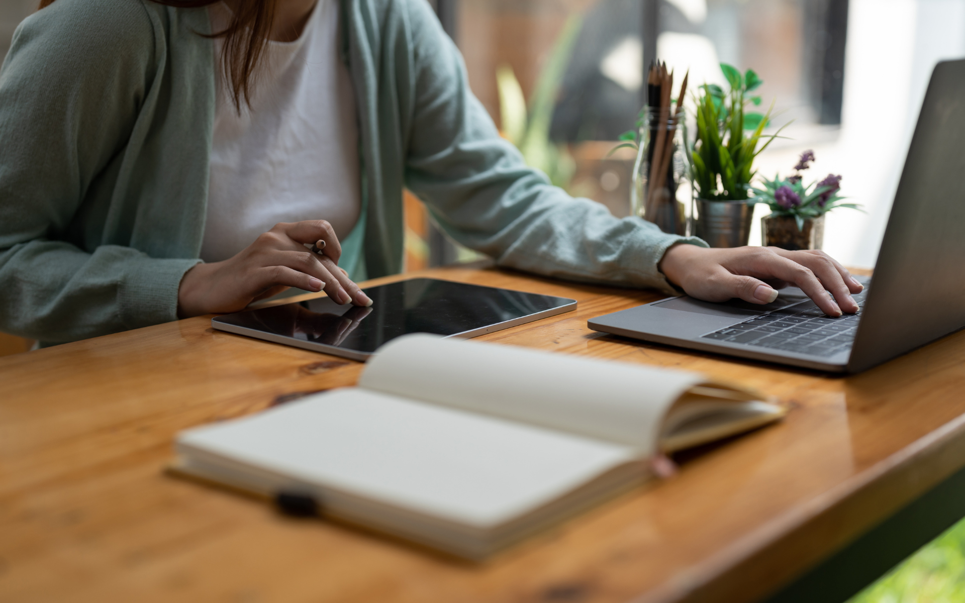 A person in a light green sweater works at a wooden desk with a laptop, a tablet, and an open notebook.