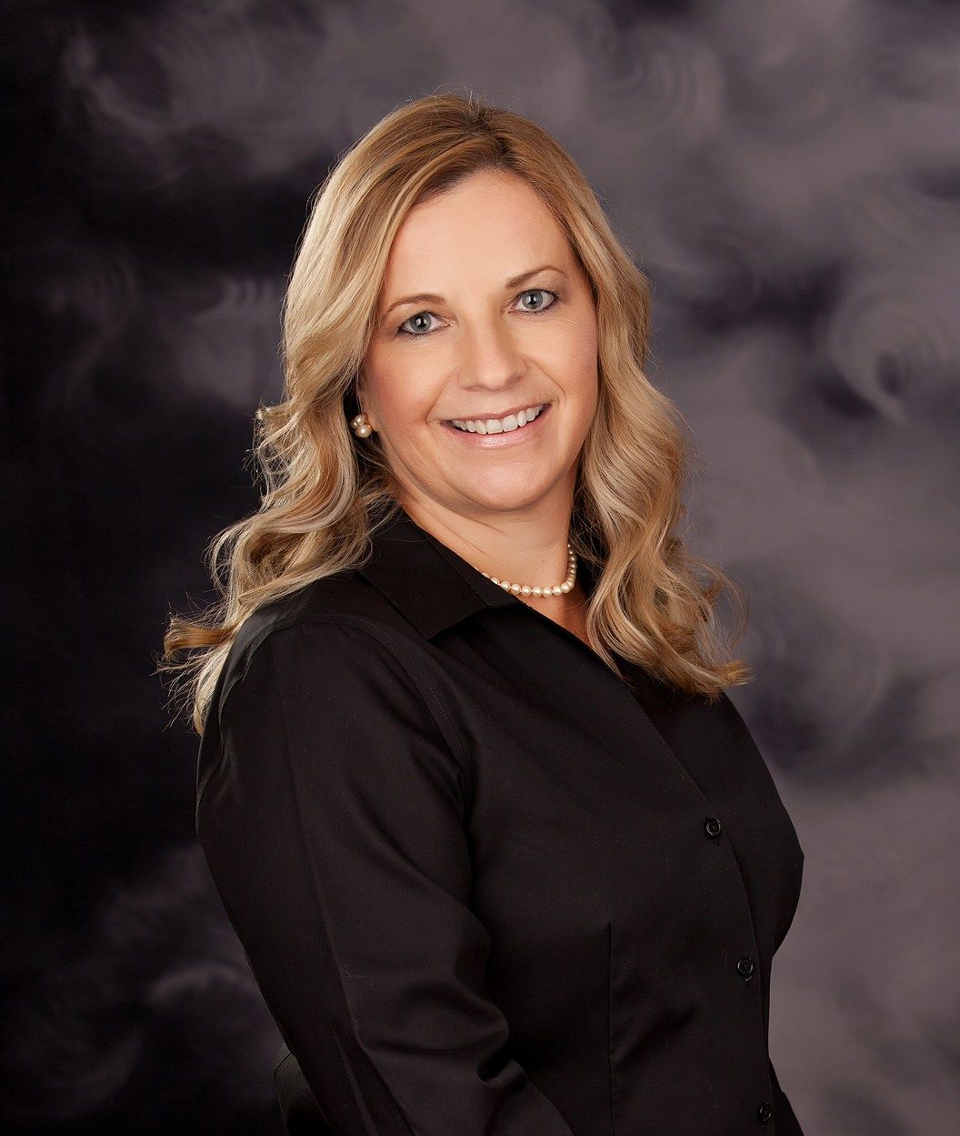 A professional headshot of a smiling woman with blonde hair, wearing a black shirt and a pearl necklace against a dark background.