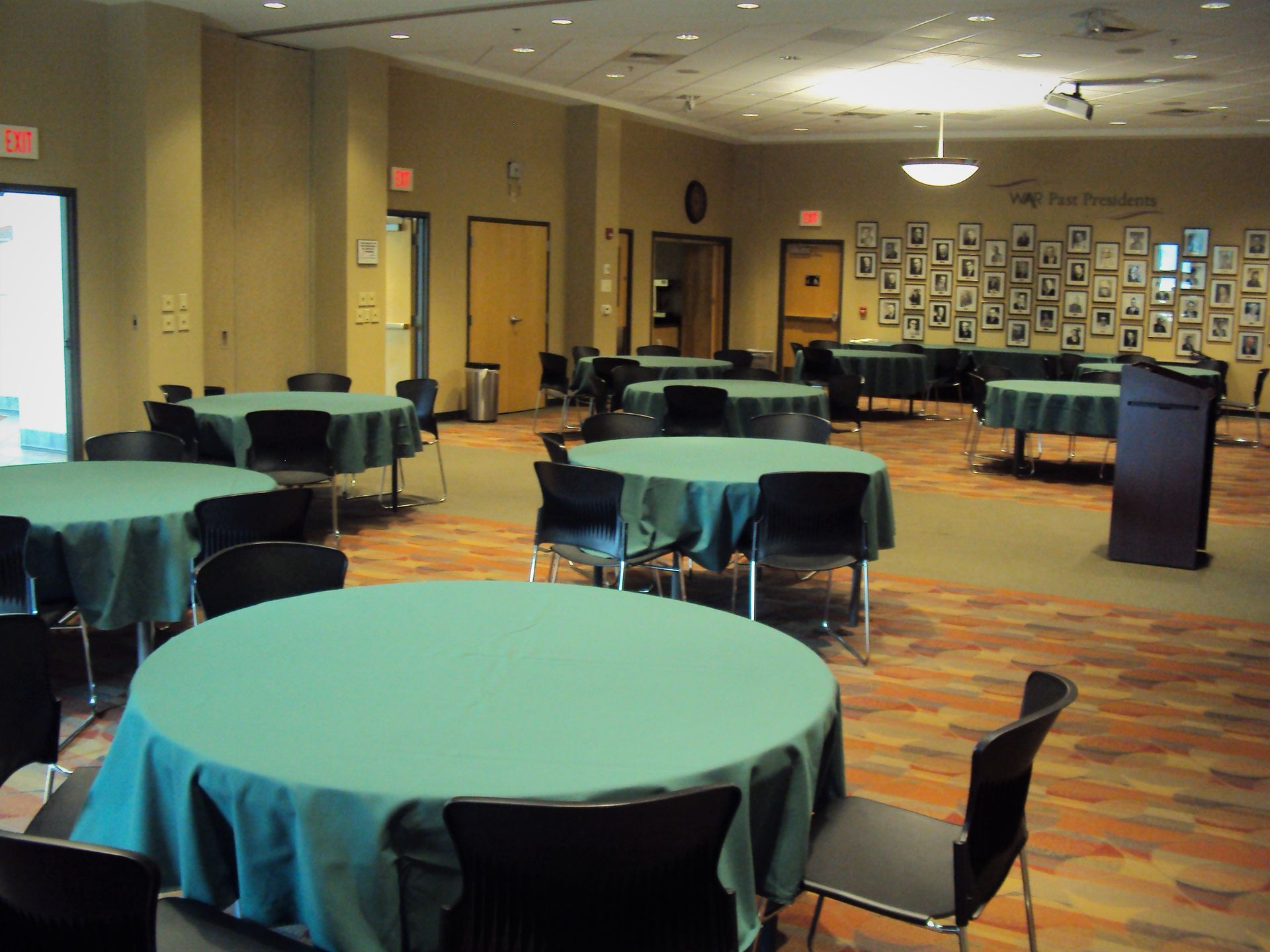 A banquet hall with round tables covered in green cloths, black chairs, and a display of framed photos on the back wall.