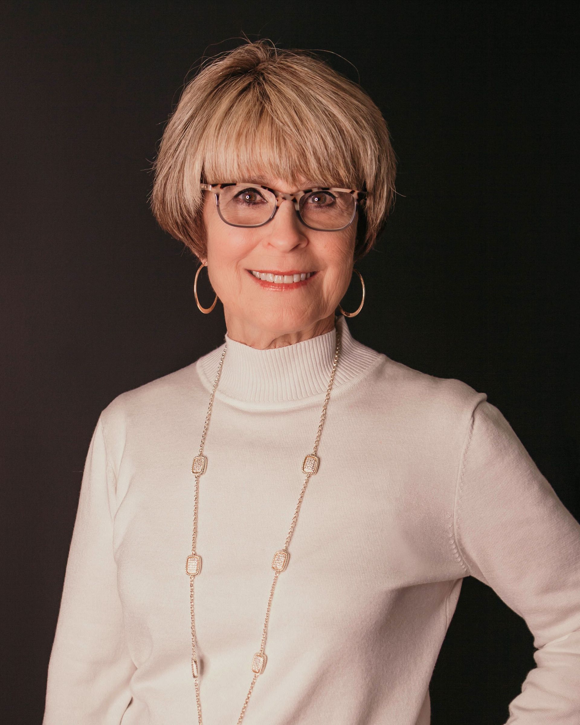 A smiling person with short, light hair wearing glasses, a white high-neck sweater, and a long necklace against black.