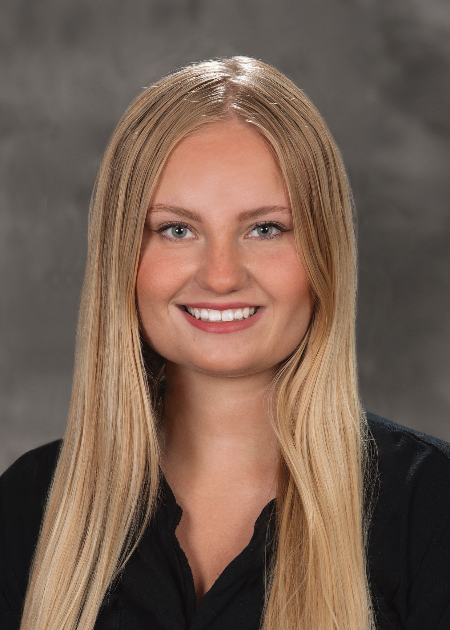 A smiling woman with curly blonde hair wearing a blue top and a light-colored patterned scarf against a dark background.