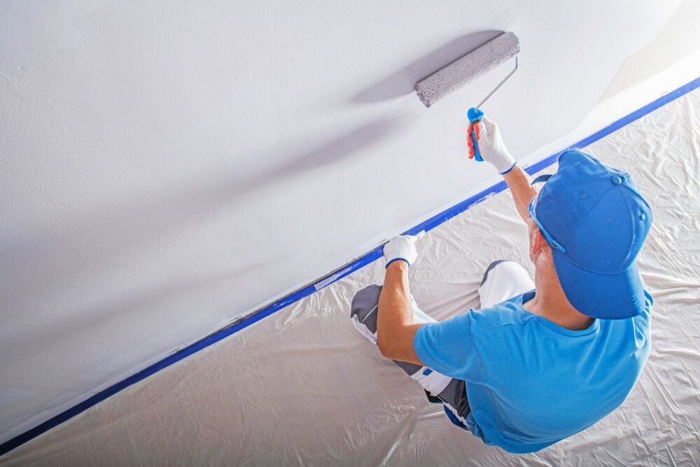 A Man Is Sitting On The Floor Painting A Wall With A Paint Roller — L & S Wirth Painting in Dubbo, NSW