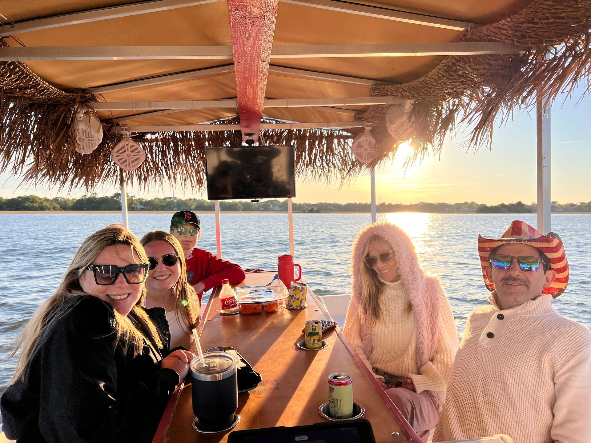 Friends seated at a tiki-style boat table on the water at sunset, smiling with drinks.