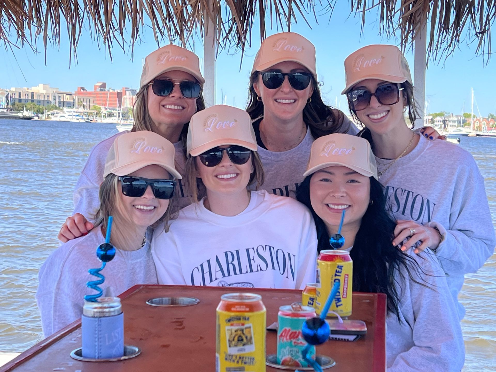 Five smiling friends in matching caps and sunglasses at a waterfront table, with Charleston shirts and drinks