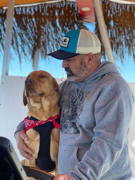 Man holding a brown dog in a red bandana under a thatched beach shelter