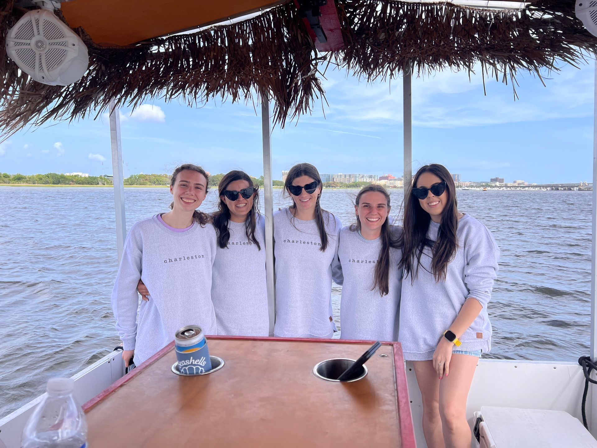 Five friends in white outfits pose on a boat with water and shoreline behind them.